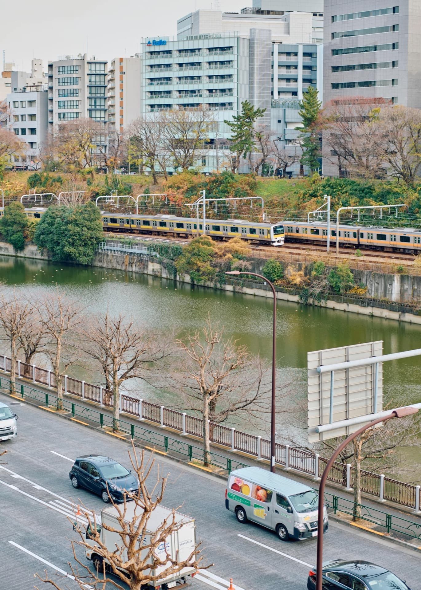 武蔵野美術大学市ヶ谷キャンパス付近の川沿いを走る電車と周囲のビル群